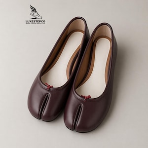 May include: A pair of dark burgundy split-toe ballet flats featuring a smooth exterior, a white interior lining, and a small red decorative knot at the center of the vamp. The logo LUXESTEPCO WHERE COMFORT MEETS CLASS is visible in the top left corner.