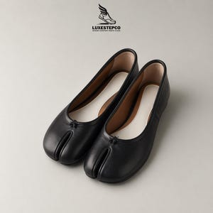 May include: A pair of black leather split-toe ballet flats. The shoes have a unique design with a separate compartment for the big toe. The interior is white, and the sole is black. The logo "LUXESTEPCO" is visible in the upper right corner.