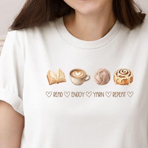 May include: A white short-sleeved t-shirt featuring a graphic print of an open book, a coffee cup with heart latte art, a ball of yarn, and a cinnamon roll. Below the icons, the text reads: READ HEART ENJOY HEART YARN HEART REPEAT HEART.