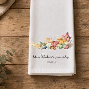 May include: A white kitchen towel featuring a watercolor illustration of various pumpkins, gourds, and autumn leaves. Below the illustration, the text reads "the Baker family" in a script font, followed by the text "est. 2016" in a simple font.