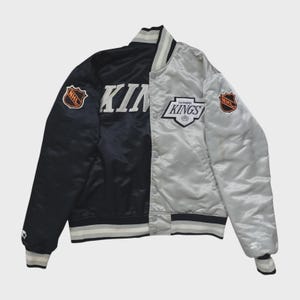 Op de afbeelding: Een tweekleurig satijnen bomberjack met een zwarte linkerkant en een zilveren rechterkant. Het jack toont de tekst KINGS, het logo van de Los Angeles Kings en NHL-patches op beide mouwen. Het heeft een gestreepte kraag, boorden en zoom.