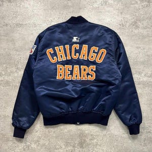 以下が含まれることがあります： 背面にオレンジ色の刺繍でCHICAGO BEARSの文字が入ったネイビーブルーのサテン製ボンバージャケット。ジャケットの上部中央にはStarterのロゴがあり、左袖には小さなNFLのワッペンが付いています。袖口はリブ編みになっています。