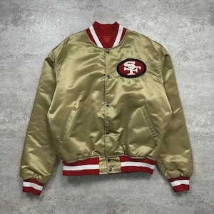 Puede incluir: Una chaqueta universitaria de satén dorado con cuello, puños y dobladillo acanalados a rayas rojas y blancas. La parte delantera tiene un cierre de botones a presión y un parche con el logo de los San Francisco 49ers en el pecho izquierdo con bordado rojo, blanco y negro.