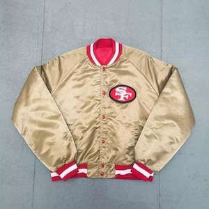 May include: A gold satin bomber jacket featuring a red and white striped collar and cuffs. The jacket has a front button closure and a red oval patch on the left chest with the white text SF. The garment has long sleeves and a ribbed hem.