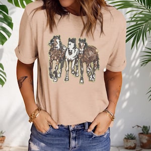May include: A beige short-sleeved t-shirt featuring a central graphic of three horses rendered in brown, white, and black sequins. The horses are depicted in a standing position, and the shirt has a crew neckline and rolled sleeve cuffs.