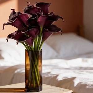May include: A bouquet of deep purple calla lilies with long green stems arranged in a tall, cylindrical amber glass vase. The flowers feature curved, trumpet-shaped petals and are positioned upright, showcasing their dark, rich color and smooth texture.
