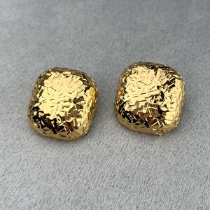 May include: A pair of gold-colored, square-shaped earrings. The earrings have a textured, hammered metal appearance. The earrings are displayed on a gray surface. These earrings are a fashion accessory.