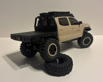 May include: A beige scale model pickup truck with a black flatbed, black roof rack, and large off-road tires. The truck is positioned with one rear wheel resting on an extra loose black tire, showcasing its detailed tread and suspension.