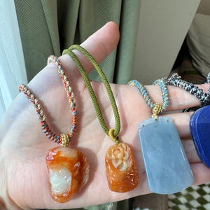 May include: A collection of gemstone pendants on colorful cords. The pendants include a monkey, a carved flower, and a rectangular blue-green stone. The cords are in shades of green, orange, and blue.