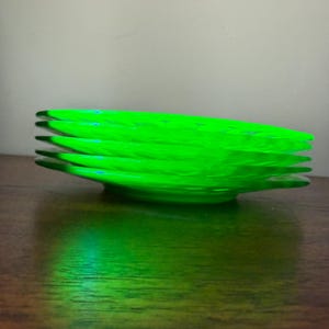 May include: A stack of five translucent, vibrant neon green glass plates. The plates feature a circular, ridged pattern radiating from the center toward the scalloped edges, creating a distinct texture that catches the light.