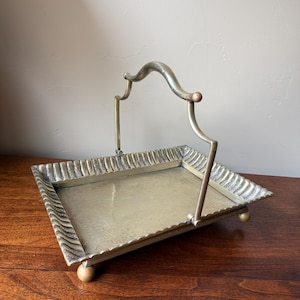 May include: A rectangular brass serving tray with a metallic finish, featuring a raised, textured border with a wavy pattern. The tray includes a central arched handle and stands on four small, rounded ball feet at each corner of the base.