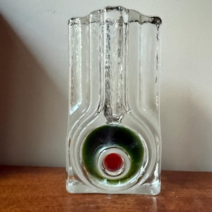 May include: A clear glass vase with a rectangular silhouette and vertical fluted channels. The center features a circular indentation containing a deep green glass orb with a small, solid red glass sphere embedded in the very center of the design.