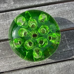 May include: A circular, transparent, green glass flower frog with multiple holes for arranging flowers. The glass has a textured surface and a bright green hue. The flower frog is designed to hold flower stems in place.