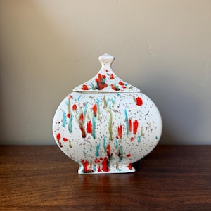 May include: A white ceramic lidded jar with a rounded, flattened body and a pointed finial on the lid. The surface features a speckled pattern with vertical streaks of bright red, turquoise, and dark brown paint splatters across the exterior.