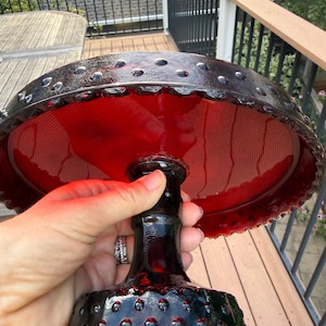 May include: A deep red glass cake stand featuring a circular plate and a pedestal base. The rim of the plate and the base are decorated with a raised hobnail pattern. The glass is translucent, allowing light to pass through the vibrant red color.