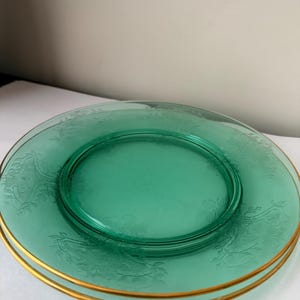 May include: Two stacked round plates made of translucent green glass. Each plate features a delicate embossed floral pattern around the rim and a thin, metallic gold band along the outer edge, creating a refined and classic appearance.