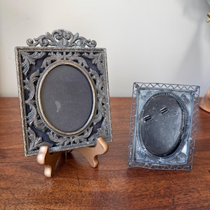 May include: Two rectangular picture frames with oval openings. The larger frame features an ornate, dark metallic finish with intricate scrollwork and sits on a small stand. The smaller frame has a delicate, open-work wire border design.