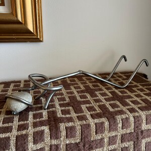 May include: A handcrafted metal sculpture featuring a stone wrapped in wire, connected to a long, curved metal arm with two upward-curving hooks. The sculpture rests on a brown patterned surface.
