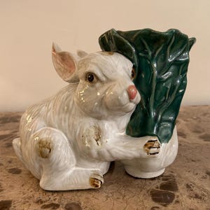 May include: A ceramic figurine of a white rabbit holding a green leafy vegetable. The rabbit has pink ears and nose, with brown accents on its paws and body. The figurine is a decorative item.