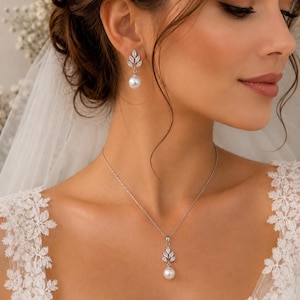 May include: A silver necklace and matching earrings set featuring a leaf design with clear crystals and a dangling white pearl. The jewelry is worn with a white lace dress and veil, suitable for a wedding or special occasion.