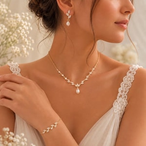 May include: A gold-toned jewelry set consisting of a necklace, a bracelet, and drop earrings. Each piece features a delicate leaf-shaped design set with clear stones and a single white teardrop pearl pendant on the necklace and earrings.