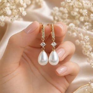 May include: A pair of gold-toned drop earrings featuring a teardrop-shaped white faux pearl suspended from a small round clear cubic zirconia stone. The upper portion of the earring consists of a slender gold-toned bar encrusted with tiny clear stones.
