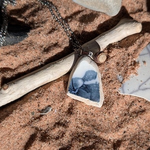 May include: A handcrafted necklace with a pendant made from a piece of ceramic, displaying a blue floral pattern. The pendant is suspended from a silver-toned chain. The necklace is displayed on sand, with driftwood and stones in the background.