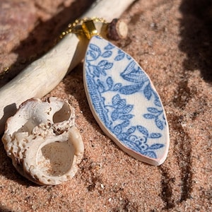 May include: A gold chain necklace with a pendant made from a piece of blue and white patterned ceramic. The pendant is oval-shaped. The necklace rests on sand with a piece of driftwood and a seashell.