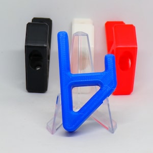 Könnte beinhalten: Ein blaues, H-förmiges 3D-gedrucktes Objekt steht im Fokus, mit einem transparenten Kunststoffständer. Weitere 3D-gedruckte Objekte in Schwarz, Weiß und Rot sind im Hintergrund. Die Objekte scheinen klein zu sein.