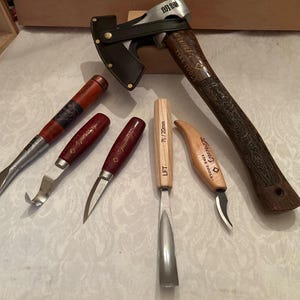 May include: A collection of woodworking tools including a hatchet with a leather sheath, a wide chisel, a curved gouge, a small carving knife, a 7L/20mm gouge, and a hook knife. The tools feature wooden handles with engraved branding and metal blades.