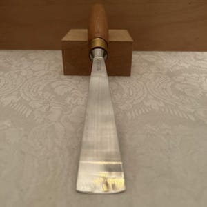 May include: A metal woodturning chisel with a long, flat, tapered blade and a smooth, cylindrical wooden handle. The tool features a brass ferrule connecting the blade to the handle, with the text ASHLEY ILES MADE IN ENGLAND engraved on the metal.