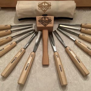 May include: A set of twelve wood carving chisels with light wood handles and metal blades, arranged in a fan shape around a central wooden mallet. Each tool is marked with LFT and a size. A canvas roll with the Lorent logo is at the top.