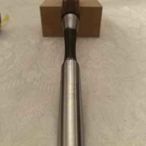 May include: A metal gouge chisel with a long, curved, polished steel blade and a dark brown wooden handle. The handle features a small, circular, gold-colored label with red Japanese characters printed on the top section of the wood.