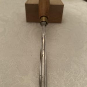 May include: A woodworking chisel with a wooden handle and a metal blade. The handle is a light brown color, and the blade is silver. The chisel is resting on a small, light brown wooden block.