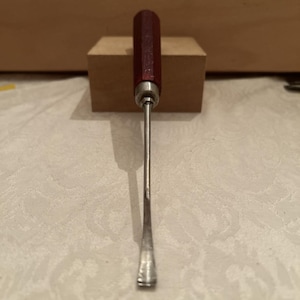 May include: A hand tool featuring a dark red, hexagonal wooden handle and a long, slender metal shaft that terminates in a curved, scoop-like blade. The metal shaft connects to the handle via a silver-colored metal ferrule at the base of the grip.