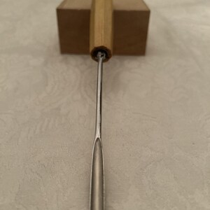 Könnte beinhalten: Ein Werkzeug aus Edelstahl und Holz mit einem Holzgriff und einer Metallklinge. Das Werkzeug ist für einen bestimmten Zweck bestimmt, wahrscheinlich in der Holzbearbeitung oder im Kunsthandwerk. Der Griff ist hellbraun und die Klinge ist silberfarben.