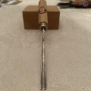 Puede incluir: Un formón para carpintería con mango de madera y hoja plateada. El mango tiene un acabado marrón claro y una etiqueta roja y blanca. La hoja es recta y se estrecha hasta un borde afilado. Un bloque de madera está en el fondo.