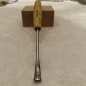 Op de afbeelding: Een metalen beitel met houten handvat en zilveren lemmet. De handgreep is lichtgekleurd hout met een cilindrische vorm. Het lemmet is een lange, slanke metalen staaf die aan het uiteinde breder wordt. Op de achtergrond staat een kleine bruine doos.