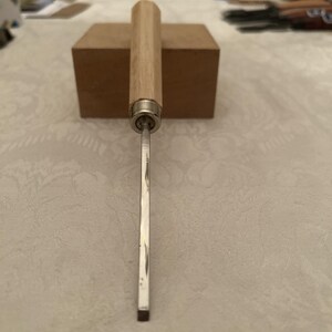 Puede incluir: Un formón para carpintería con un mango de madera clara y una hoja plateada. El formón está colocado verticalmente, con la hoja hacia abajo. El mango es cilíndrico y parece estar hecho de madera de color claro. La hoja es rectangular y tiene un borde afilado.