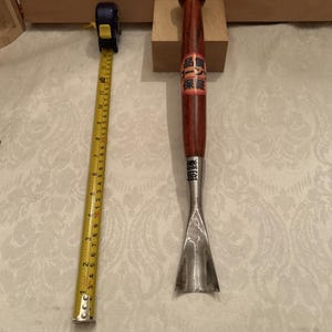 May include: A wood carving gouge with a long, dark brown wooden handle and a curved metal blade. A yellow measuring tape is placed alongside the tool, showing a length of approximately 14 inches from the base to the top of the handle.