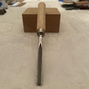 Puede incluir: Un formón para tallar madera con un mango de madera clara y una hoja plateada. El formón está colocado verticalmente, con la hoja apuntando hacia abajo. El mango es cilíndrico y parece ser de madera dura.