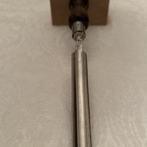 May include: A woodworking chisel with a dark wooden handle and a silver-colored blade. The chisel is positioned vertically against a light-colored background, showcasing its design and craftsmanship. The tool is designed for shaping and cutting wood.