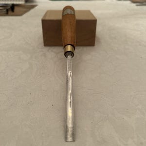 May include: A woodworking chisel with a wooden handle and a metal blade. The handle is a light brown color with a brass ferrule. The blade is silver and tapers to a sharp edge. The chisel is resting on a white surface.