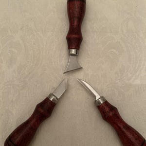 Peut inclure: Trois outils de travail du cuir sont présentés, chacun avec un manche en bois brun rougeâtre foncé et une lame en métal argenté. Les lames ont des formes différentes : une est incurvée et deux sont droites. Les outils sont disposés en triangle.