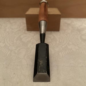 May include: A woodworking chisel featuring a wide, flat, dark-colored metal blade with a beveled edge. The tool has a cylindrical wooden handle with a silver-toned metal ferrule at the top, providing a sturdy grip for manual woodworking tasks.