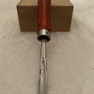 May include: A wood-handled chisel with a silver blade. The handle is a reddish-brown color with a silver ferrule. The blade has the text "DIZE MEDAL 30". The chisel is likely used for woodworking or carving.
