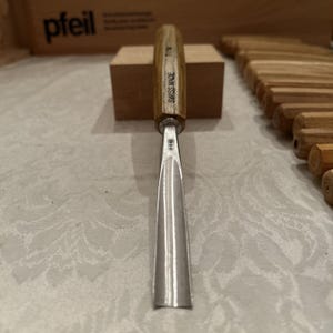 May include: A metal gouge with a wooden handle featuring the text SWISS MADE and 7/14. The tool has a curved, polished steel blade with a small engraved symbol near the bolster, positioned vertically on a light surface with other wooden handles.