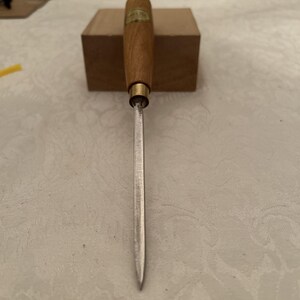 May include: A woodworking chisel with a wooden handle and a metal blade. The handle is light brown with a brass ferrule. The blade is silver and tapers to a point. The chisel is resting on a small wooden block.