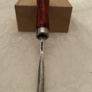 Può includere: Uno scalpello per la lavorazione del legno con un manico in legno scuro e una lama in metallo argentato. Il manico ha una parte superiore arrotondata ed è tinto di un colore marrone rossiccio scuro. La lama è di colore argento chiaro e termina a punta.