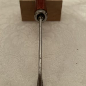 May include: A vintage metal tool with a wooden handle. The handle is a dark reddish-brown color, and the metal shaft is silver. The tool appears to be a specialized instrument, possibly for woodworking or crafts.